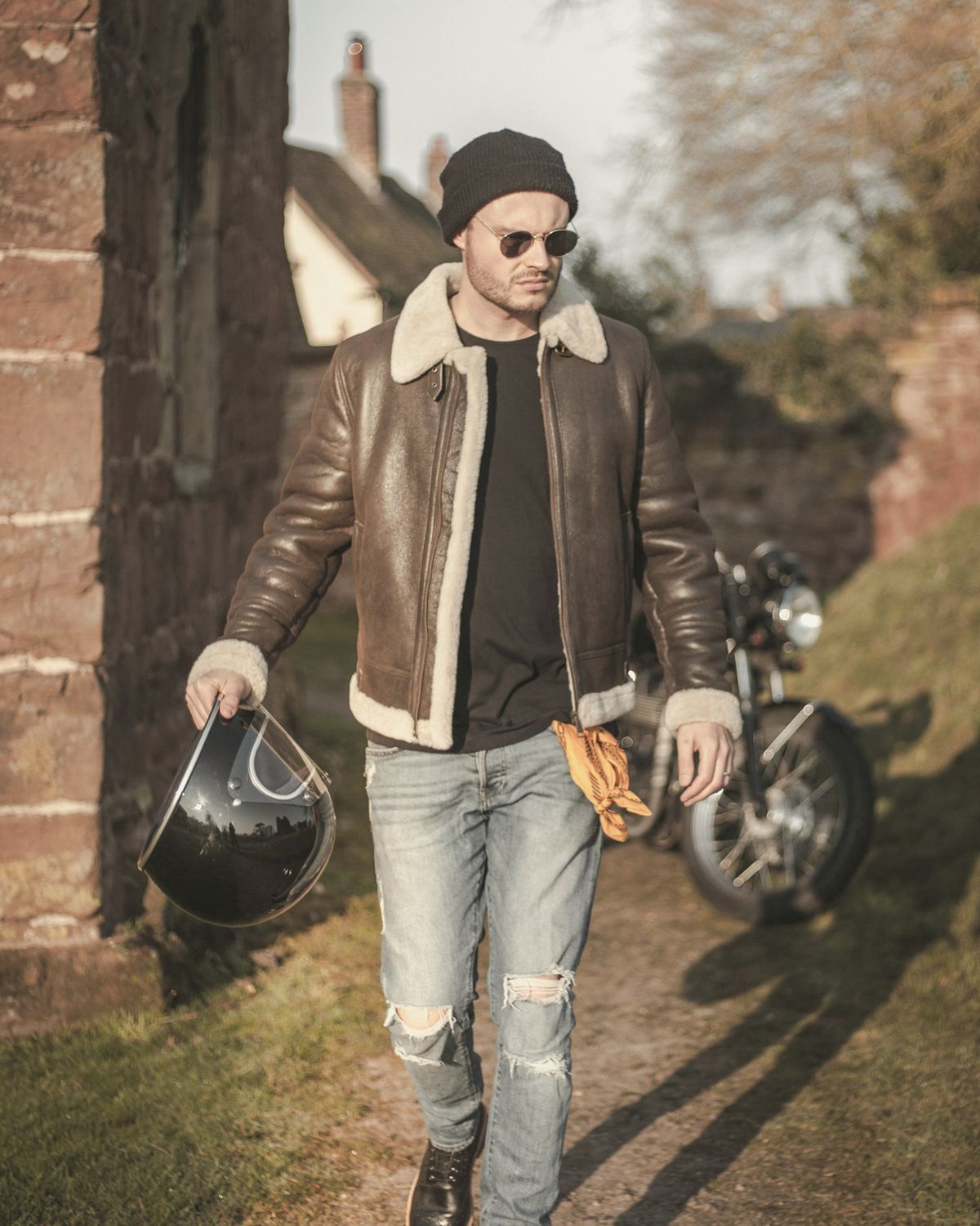 Man in a brown leather and shearling flight jacket carrying a helmet, walking past a motorbike