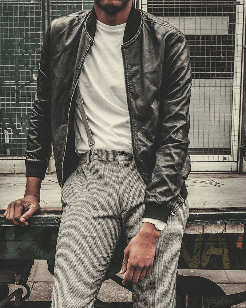 Man in a black leather bomber jacket with ribbed collar and cuffs, white tee and grey trousers