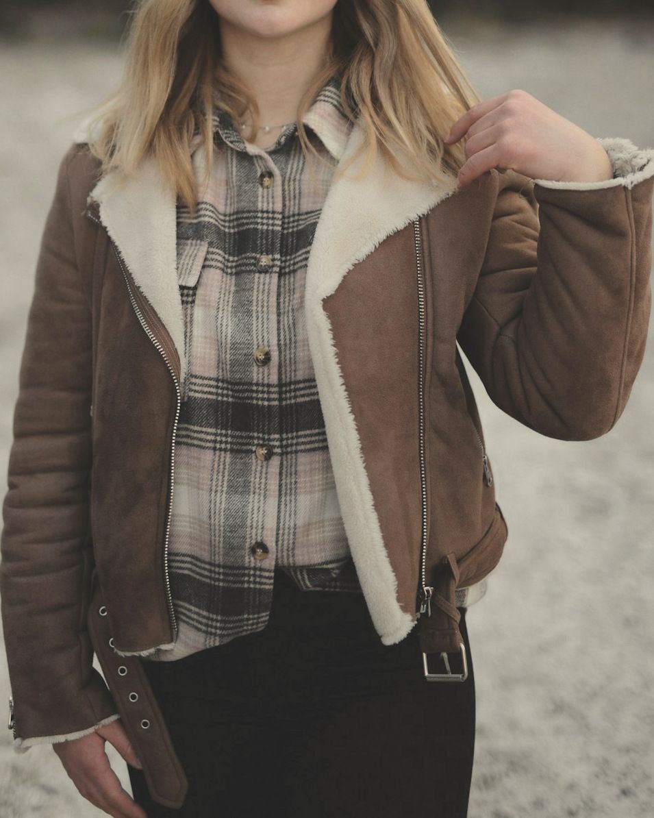 Woman in a brown suede biker jacket with a cream shearling lining, worn over a check shirt