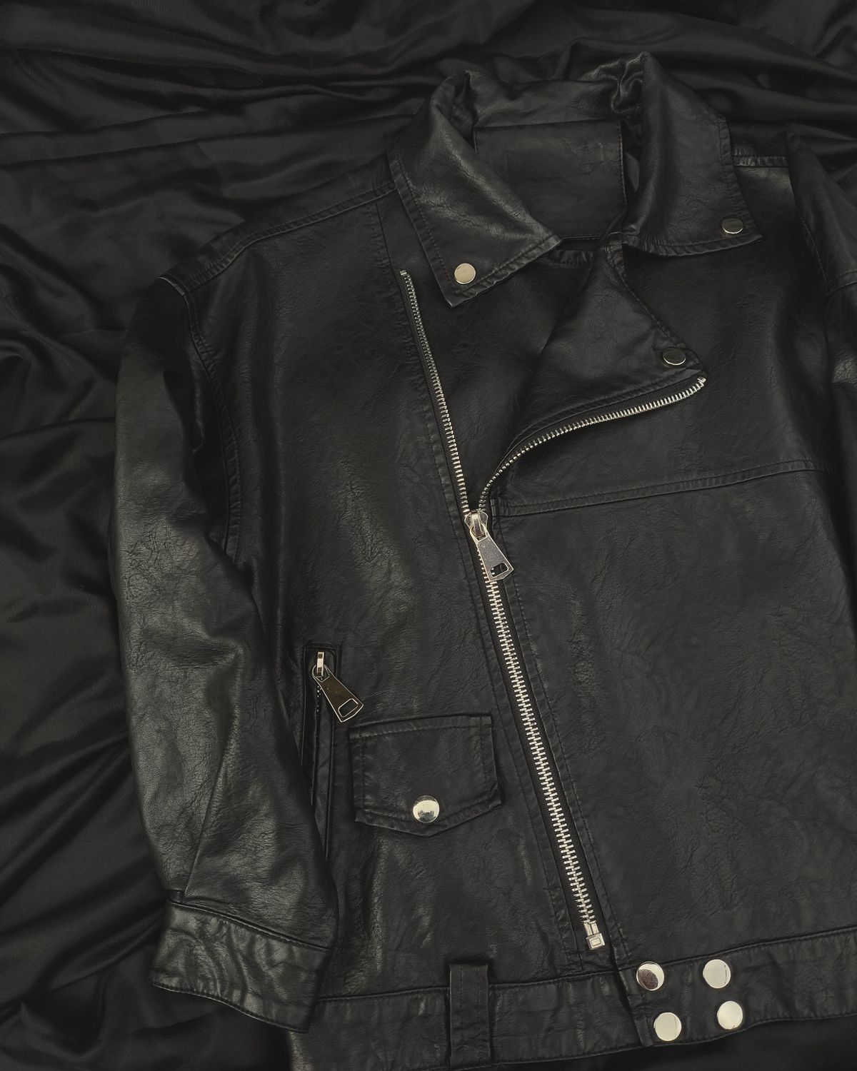 Black asymmetric biker jacket laid flat, showing the diagonal zip and snap hem