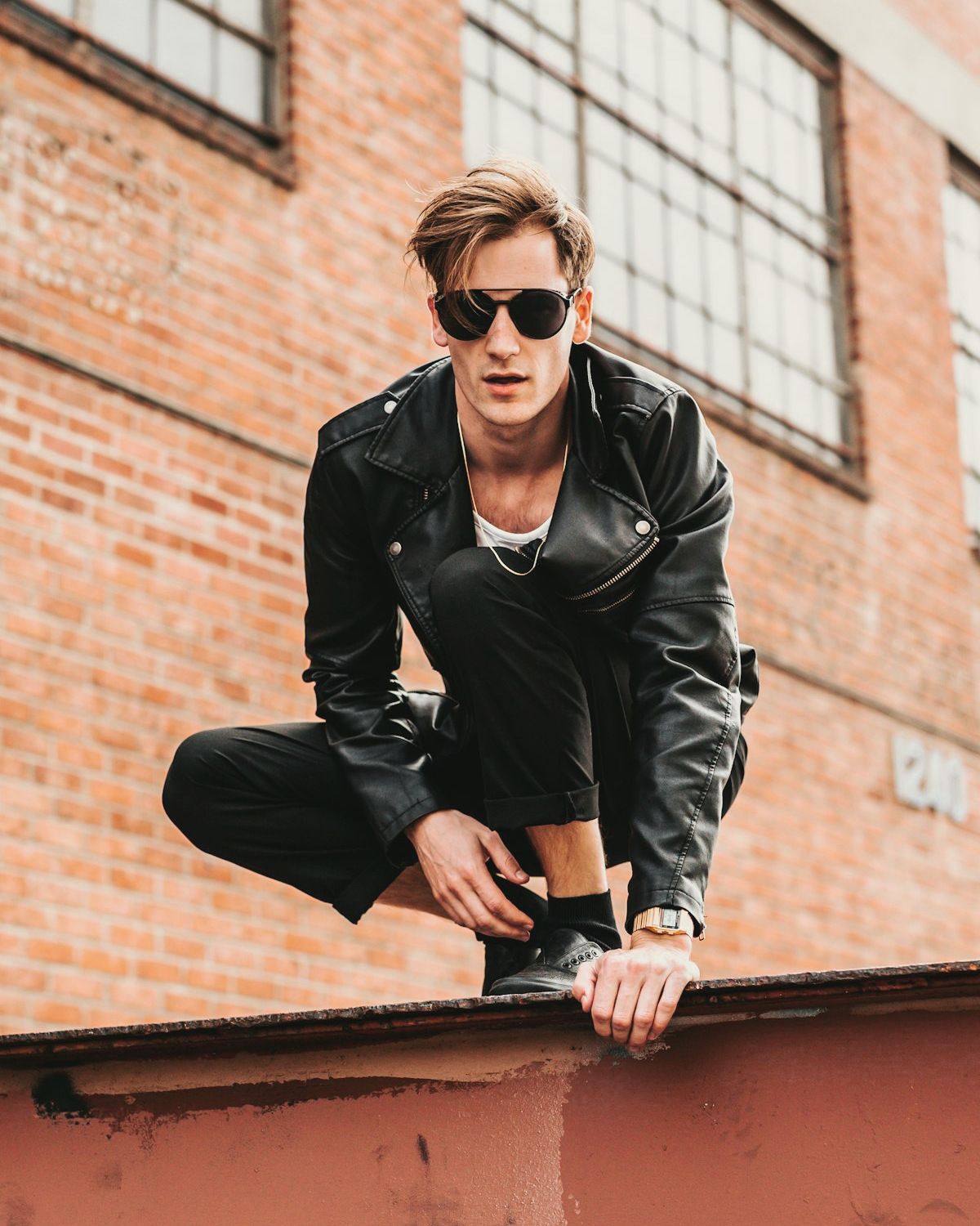 Man in a black asymmetric leather biker jacket crouching on a rusted steel beam