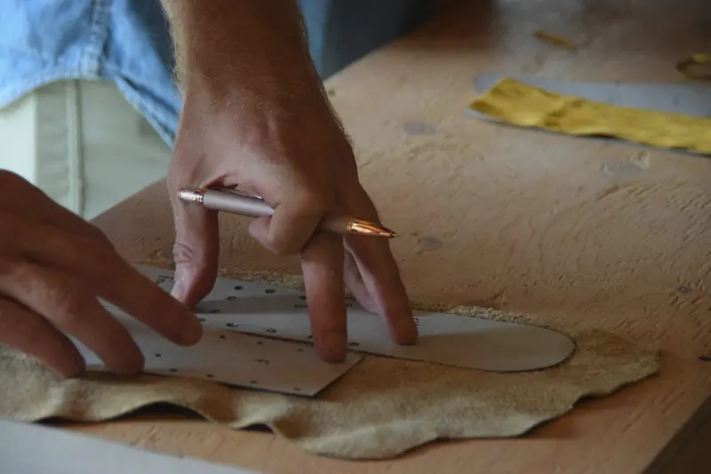 Hands marking a leather panel from a paper pattern on the workshop bench