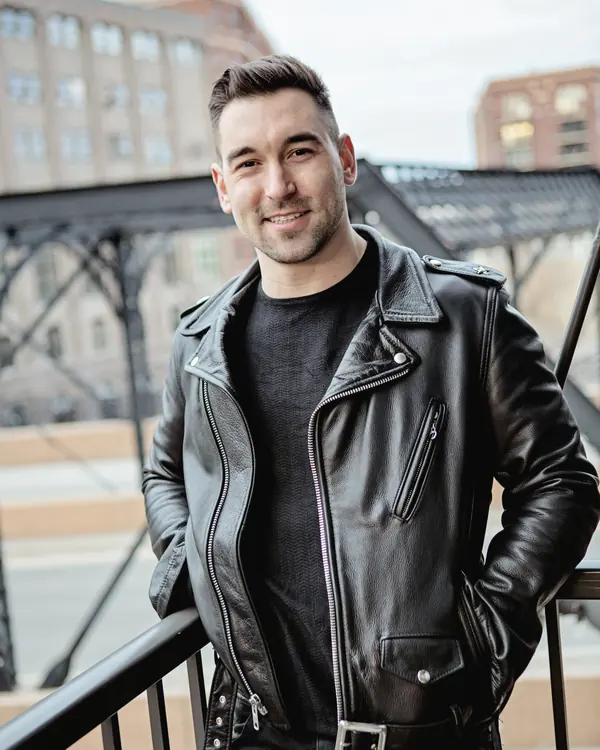 Man in a black asymmetric leather biker jacket, smiling, hands in pockets