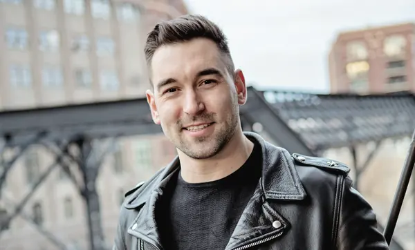 Man in a black asymmetric leather biker jacket, smiling, hands in pockets